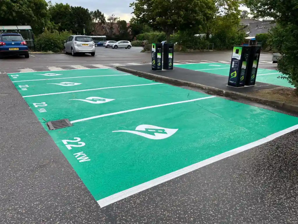 A car park features several electric vehicle charging stations with bright green painted bays, white lines, and 22 KW text on the ground, which can be described as HAZARD MARKINGS.