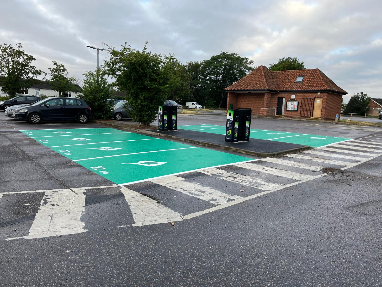 Accessible Parking Spaces A car park with several green-painted electric vehicle charging bays and two charging stations. Cars are parked in adjacent grey bays, and a small brick building is in the background.
