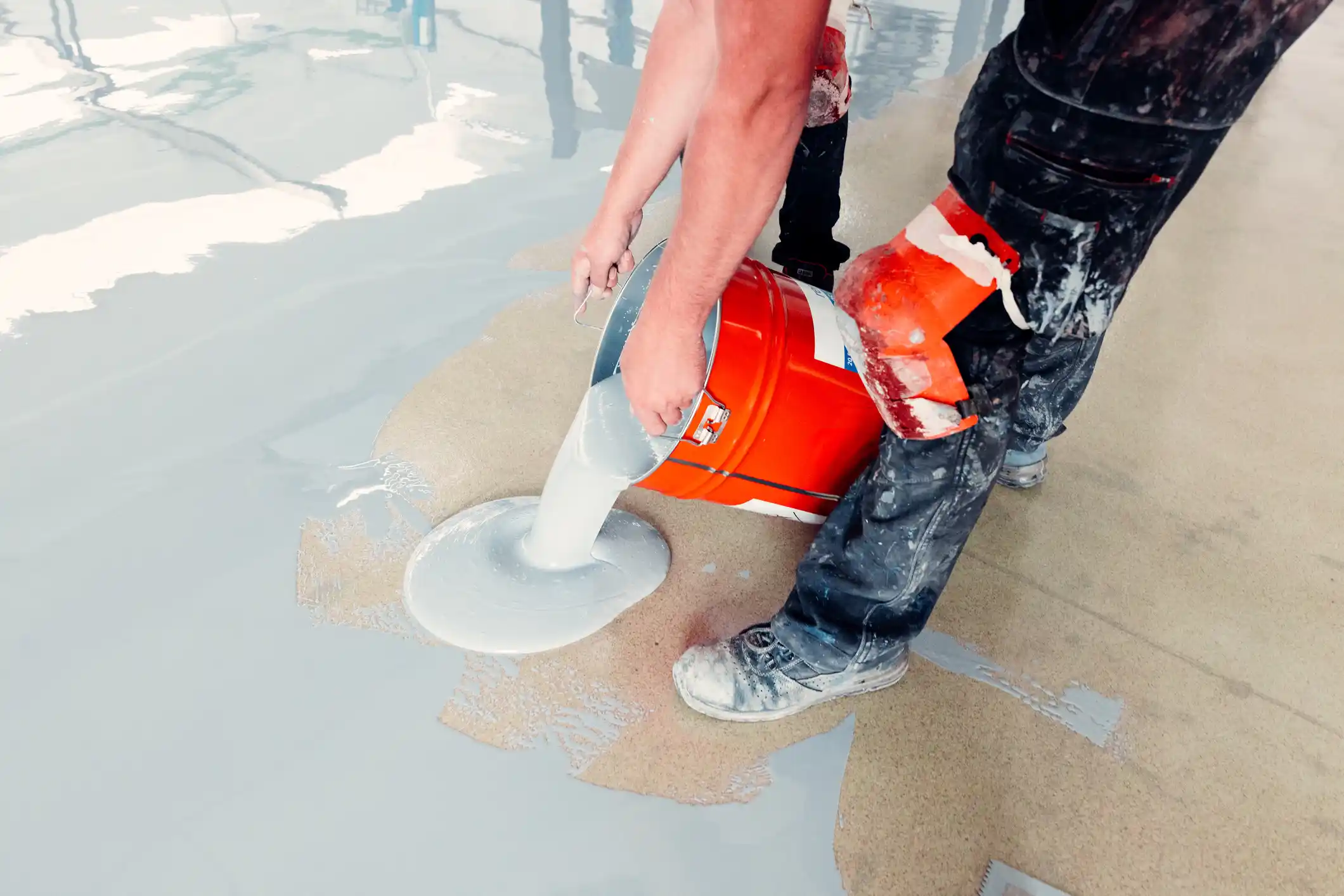 Person pouring grey epoxy floor coating from a bucket onto a concrete floor for epoxy resin floor installers